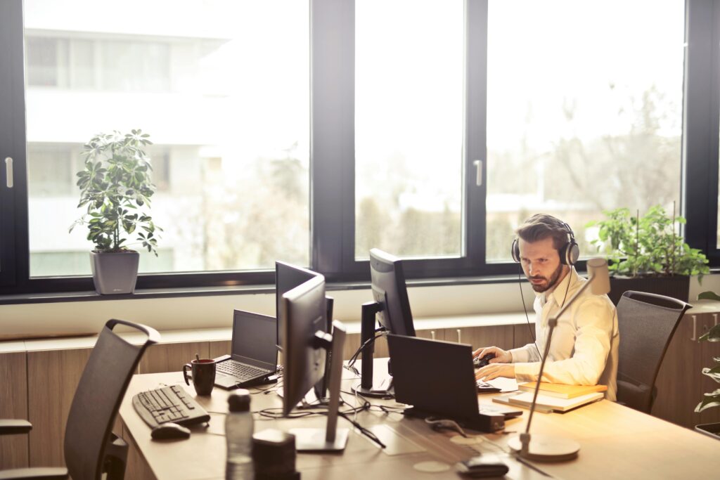 Man sitting in front of computer monitors in an office setting near bright windows.